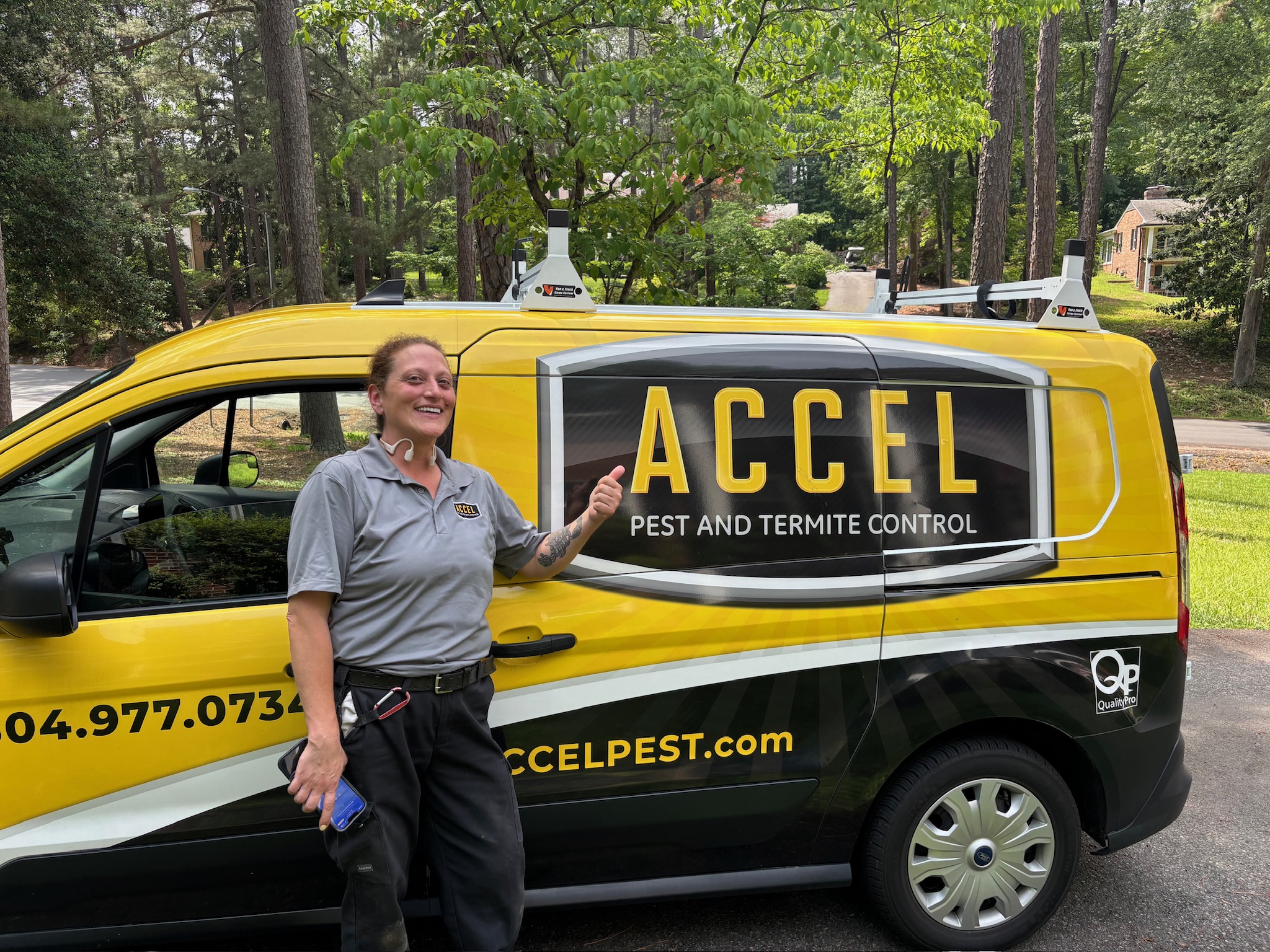 A male pest control technician wearing a grey Accel uniform kneels on a kitchen floor and shines a flashlight into a cabinet, inspecting for pests.