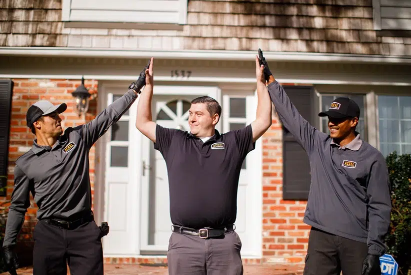 Three Accel pest control team members in dark uniforms and caps stand outside a brick house, smiling and raising their hands together in a high‑five chain.