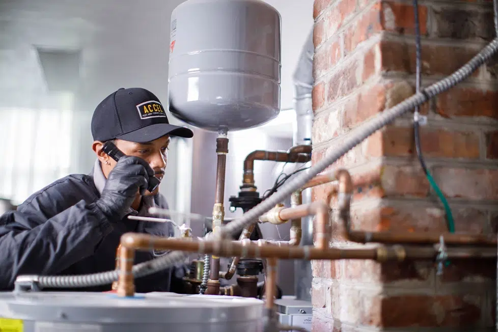 A pest control technician in a dark Accel uniform and black cap inspects pipes beside a water heater and brick wall, speaking on a phone in a utility area.