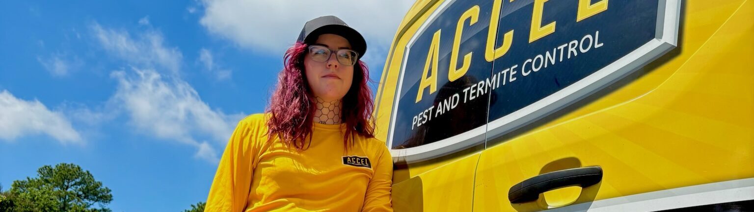 A pest control technician wearing a yellow Accel shirt stands next to a yellow service van with the Accel Pest and Termite Control logo under a blue sky.