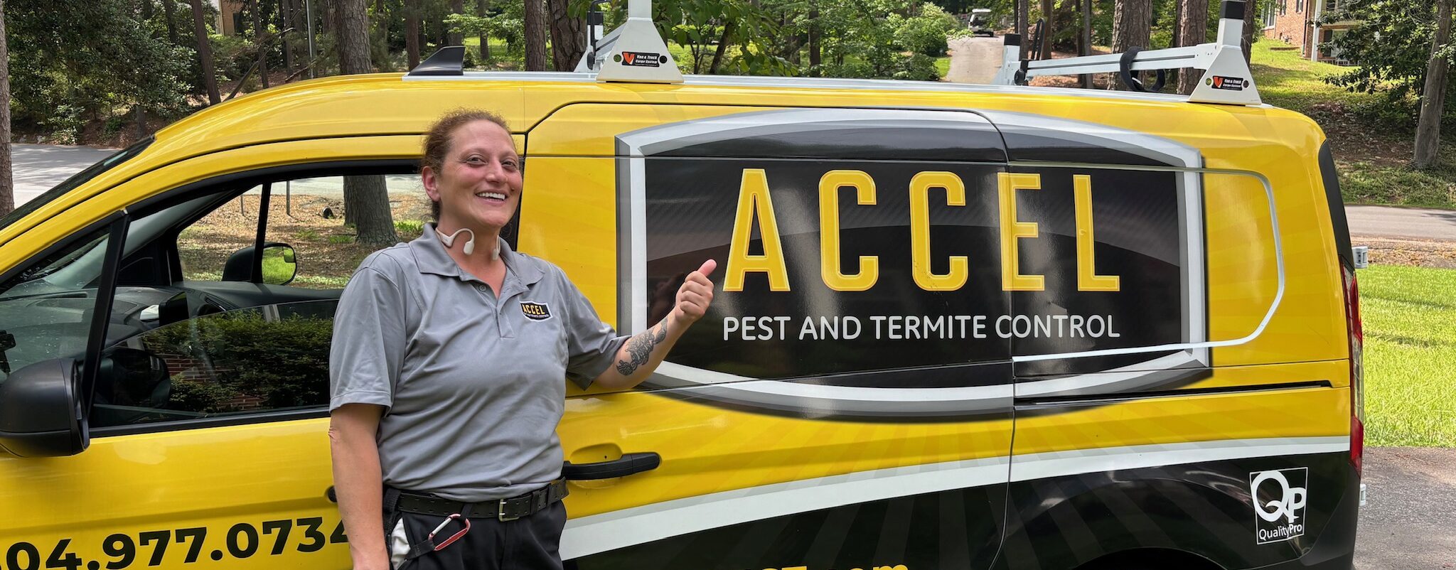 A female pest control technician wearing a grey uniform pointing to a big "Accel Pest" sign.