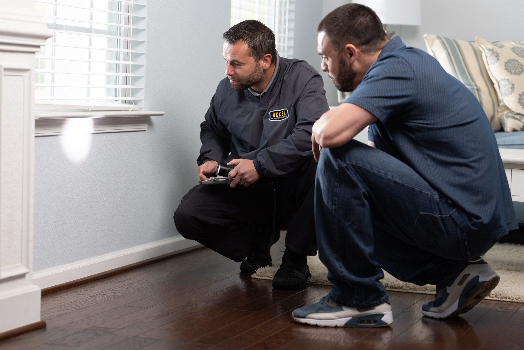 Two men observe a possible pest entry point in a living room.